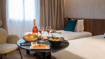 Habitación de hotel con una mesa de comedor dispuesta con comida y bebidas, una cama con un cojín azul y una ventana con cortinas.