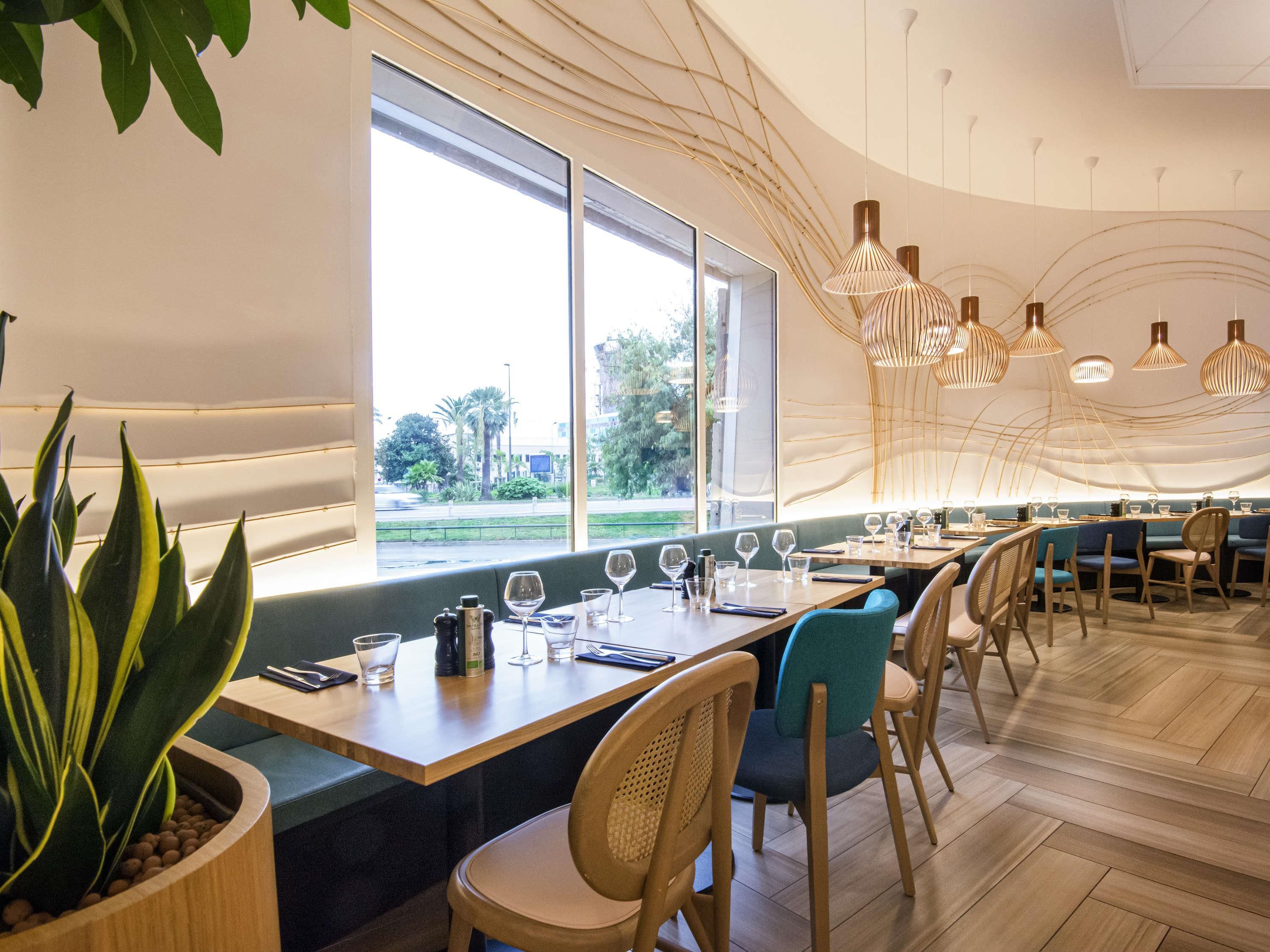 A modern restaurant interior with wooden tables, chairs, and a large window offering a view of the outdoors.