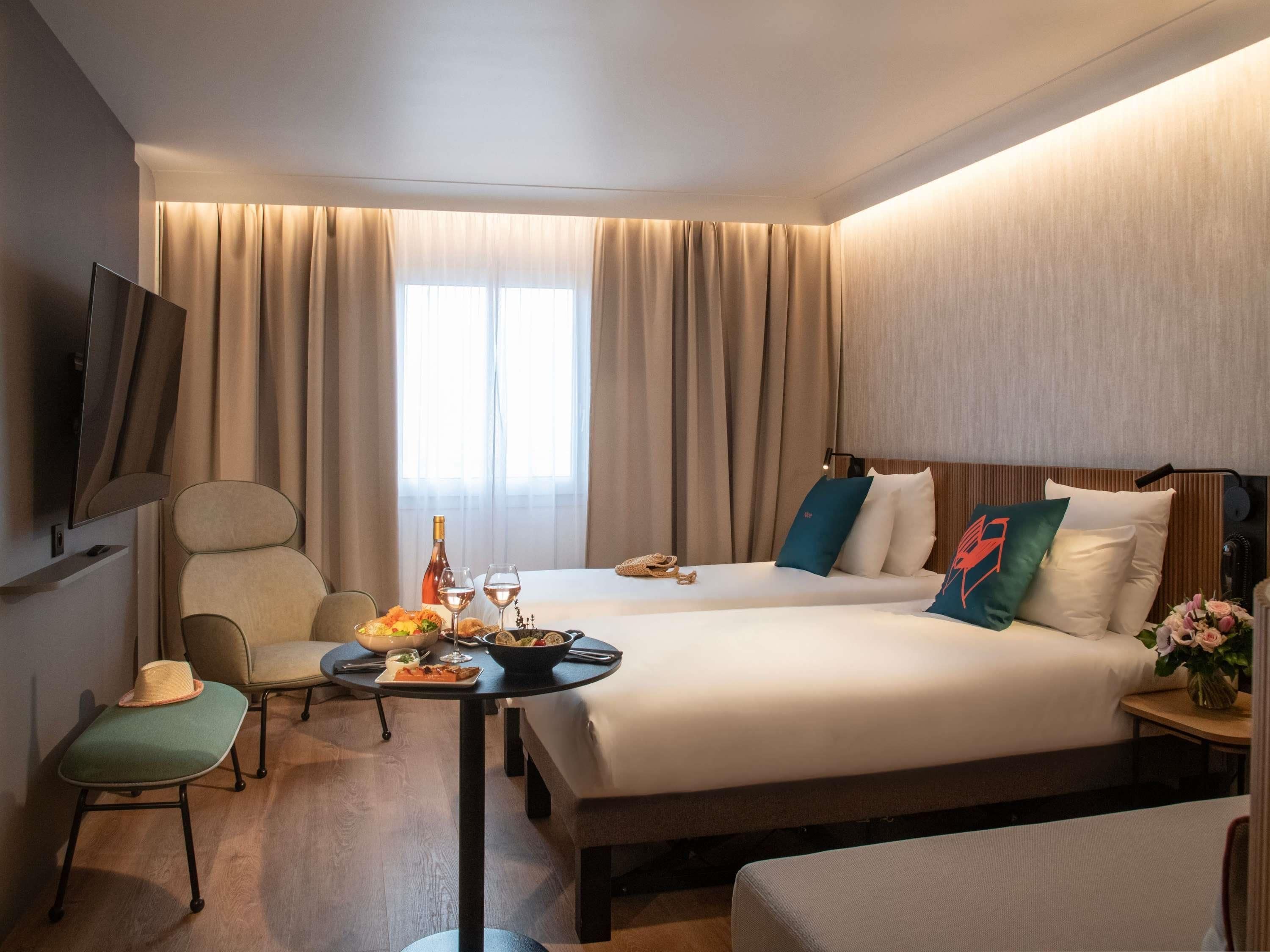 A hotel room with a bed, a desk with a chair, a TV mounted on the wall, and a window with curtains.