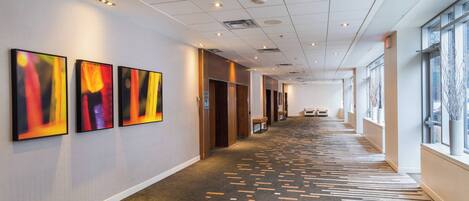 A long, well-lit hallway with a patterned carpet, large windows on one side, and abstract art on the walls.