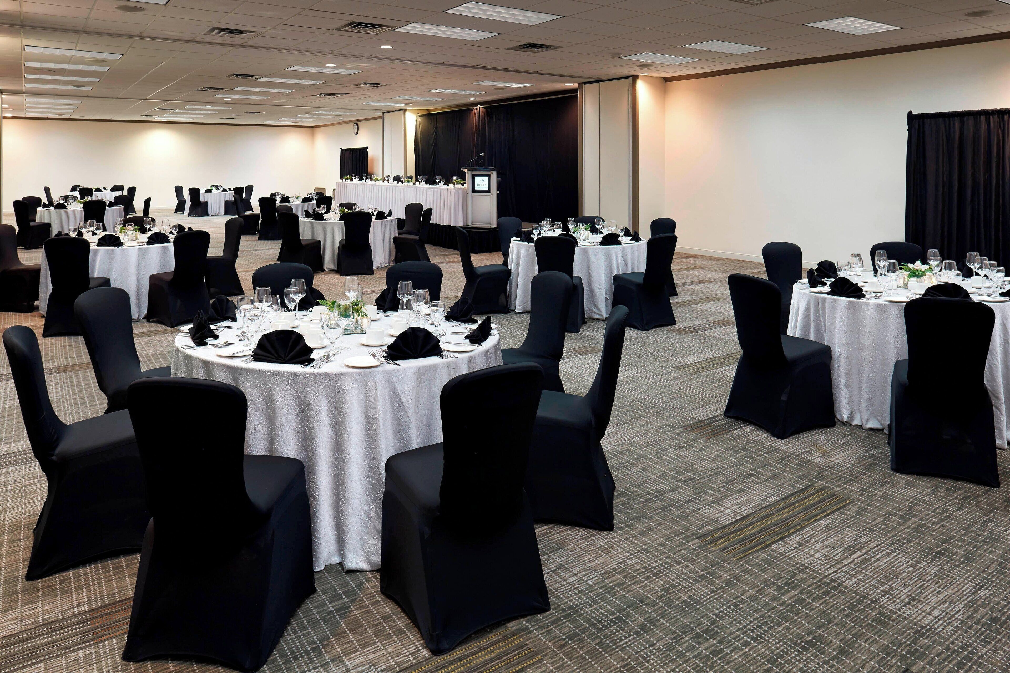 Un salón de conferencias con varias mesas redondas, cada una cubierta con mantel blanco, sillas negras y lo necesario para comer.