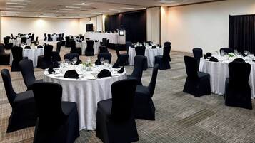 Un salón de conferencias con varias mesas redondas, cada una cubierta con mantel blanco, sillas negras y lo necesario para comer.