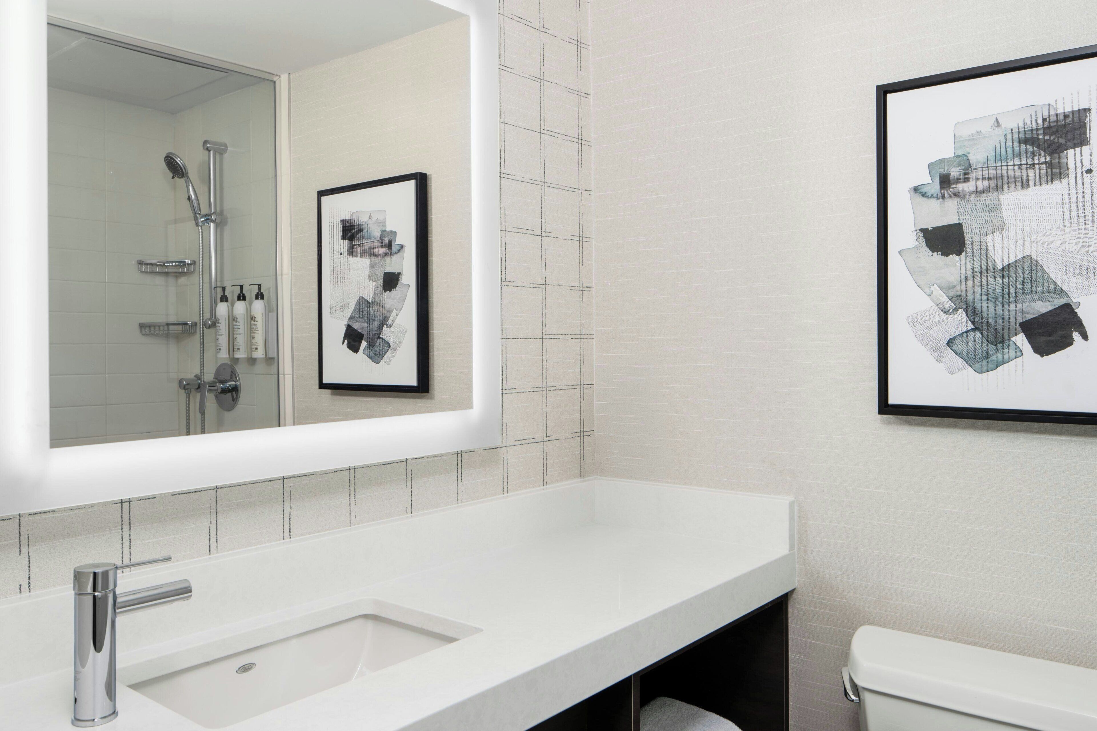 A modern bathroom with a white sink, a mirror, a shower, and two framed artworks on the wall.