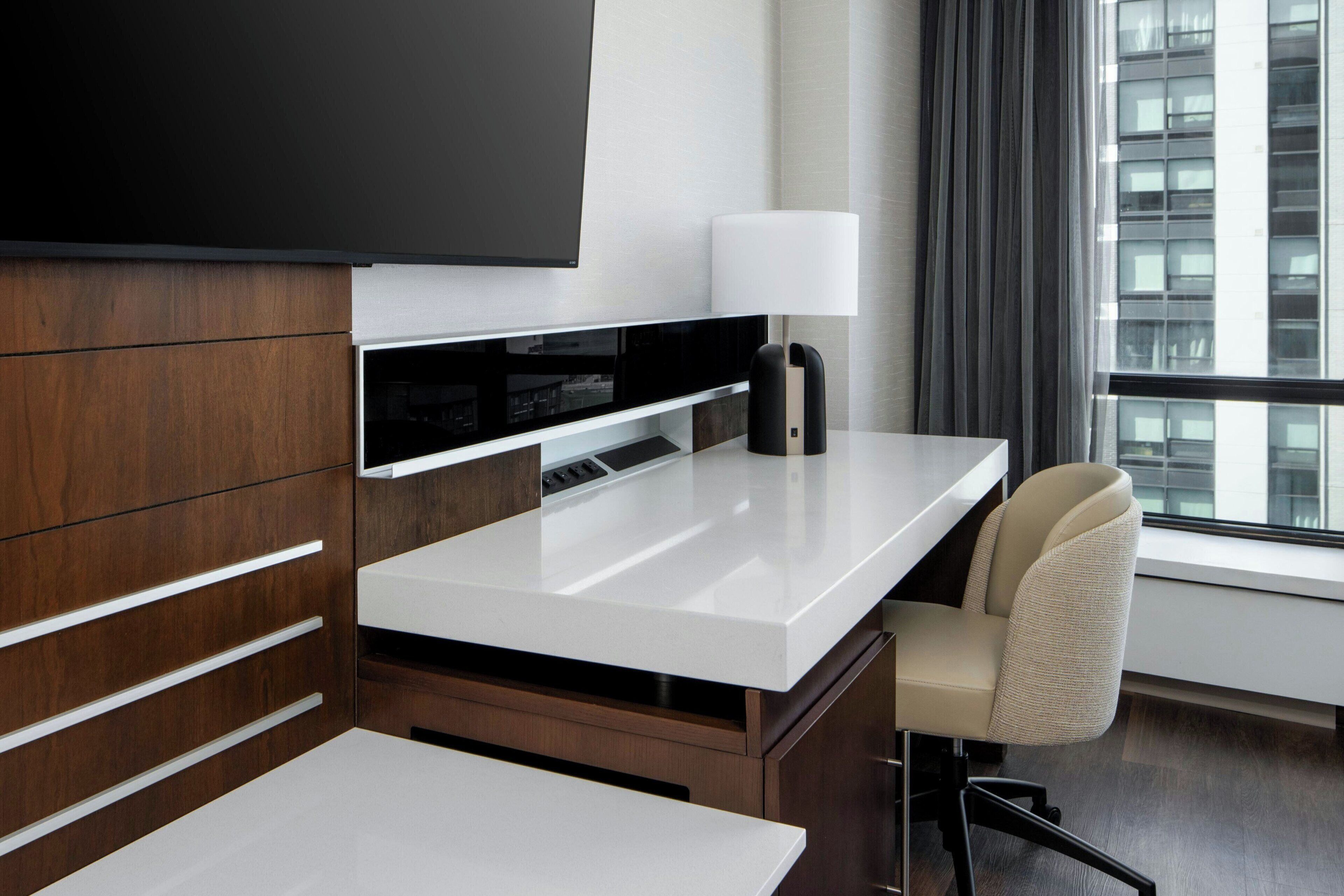 Una habitación de hotel moderna con una televisión de pantalla plana, un escritorio con una silla, una lámpara y un ventanal con vistas a la ciudad.