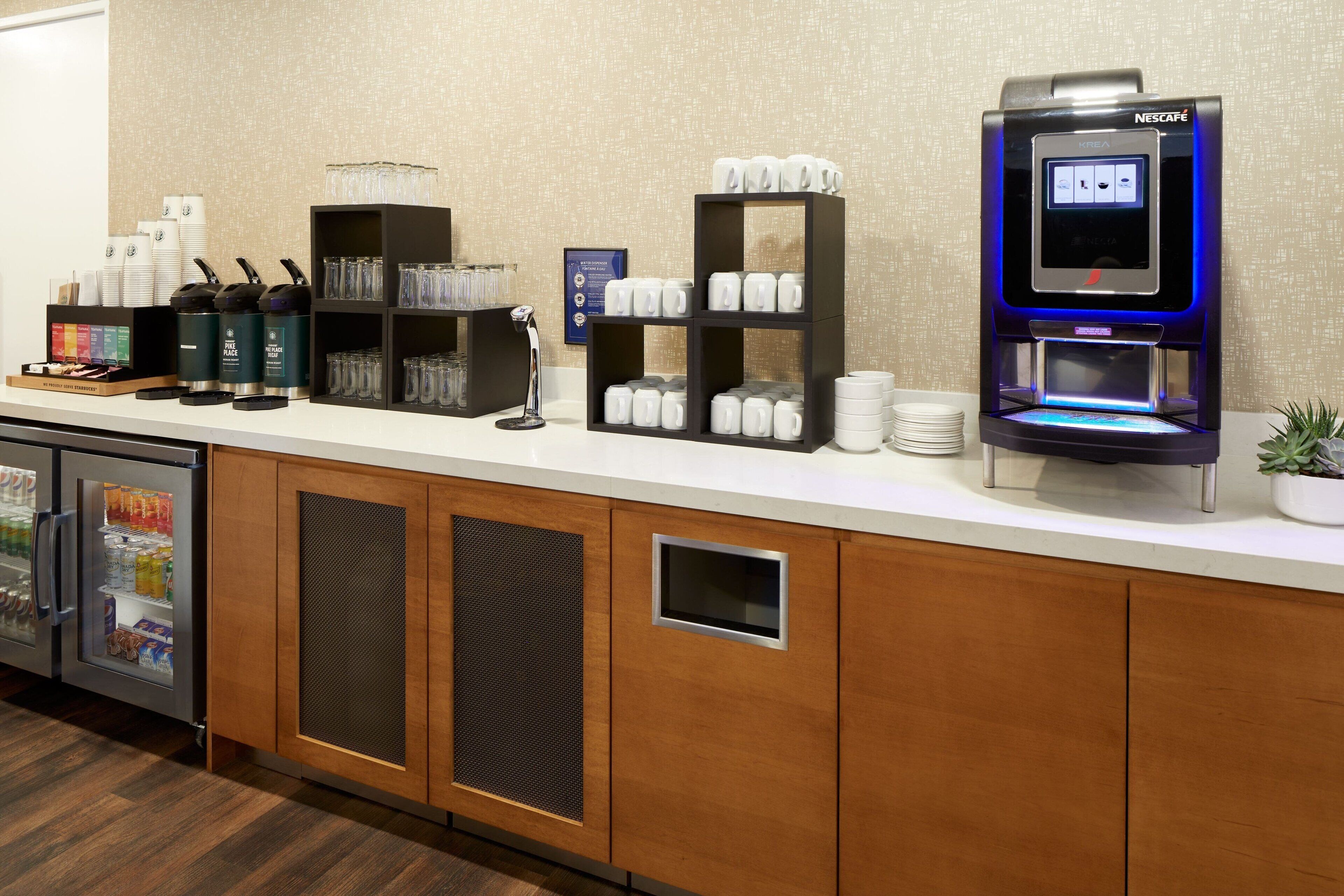 A hotel reception area with a coffee machine, beverage dispensers, and a refrigerator.