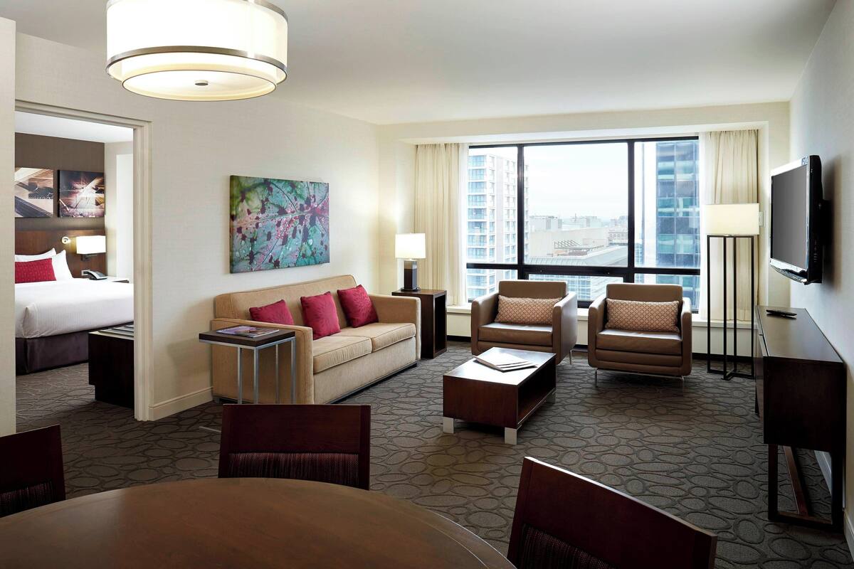 A hotel room with a bed, a sofa, two armchairs, a coffee table, a TV, and a large window with a city view.