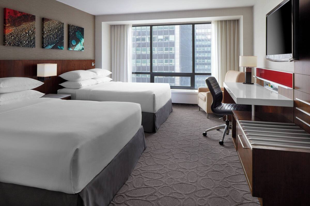 A hotel room with two beds, a desk, a chair, a TV, and artwork on the wall.
