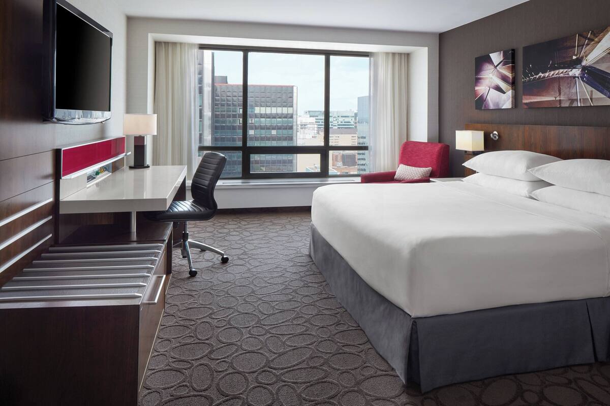 A hotel room with a bed, desk, chair, TV, and large window with a city view.