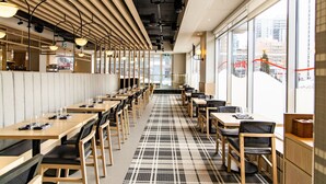 A modern restaurant with long communal tables, black and white checkered flooring, and large windows offering city views.