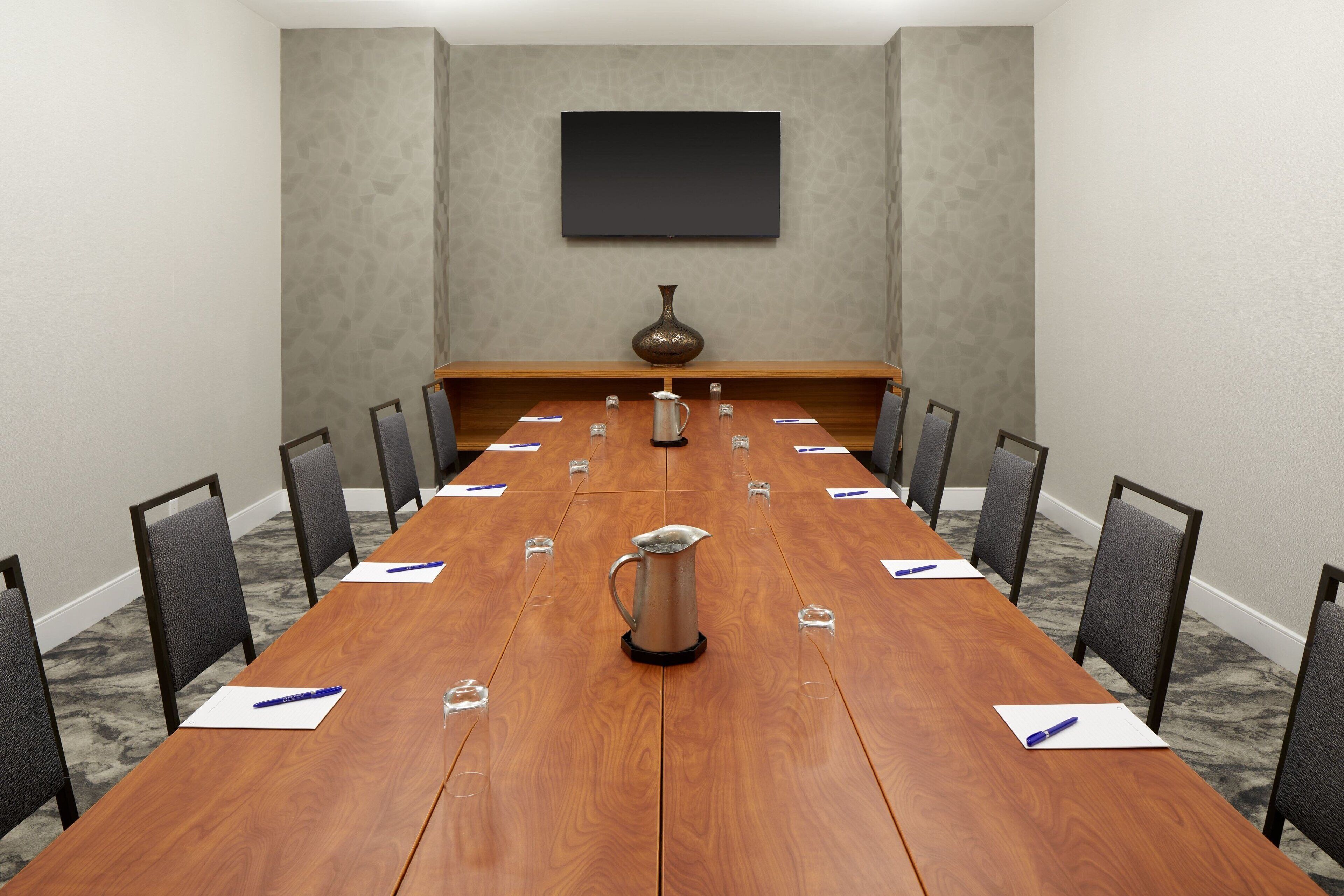 Un salón de conferencias con una larga mesa de madera, sillas, una jarra y un televisor de pantalla plana en la pared.