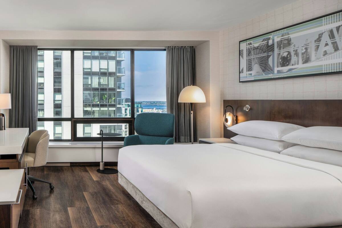 A hotel room with a large bed, a desk, a chair, a lamp, and a view of the cityscape.