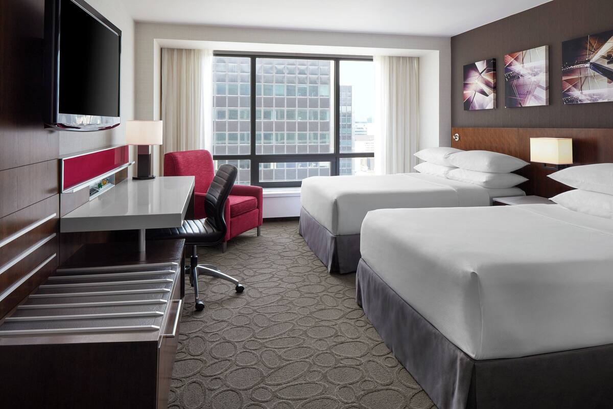 A hotel room with a bed, a desk, a chair, a TV, and a window with a city view.