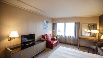 Habitación de hotel con una cama grande, un televisor en una cómoda, un sillón rojo, un escritorio con una silla y una ventana con cortinas.