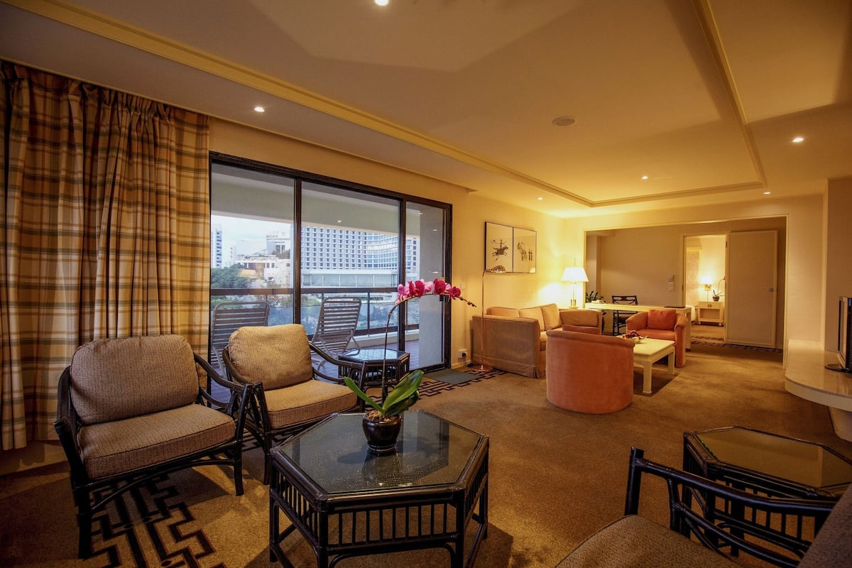 Una habitación de hotel con un ventanal grande, un balcón y una zona de estar con mesa y sillas.