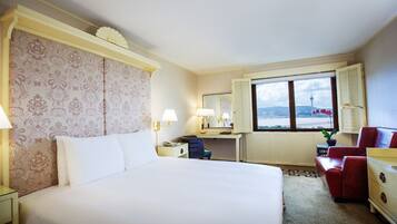 A hotel room with a large bed, a desk, a chair, and a dresser. There is a window with a view of a tower and a bridge.
