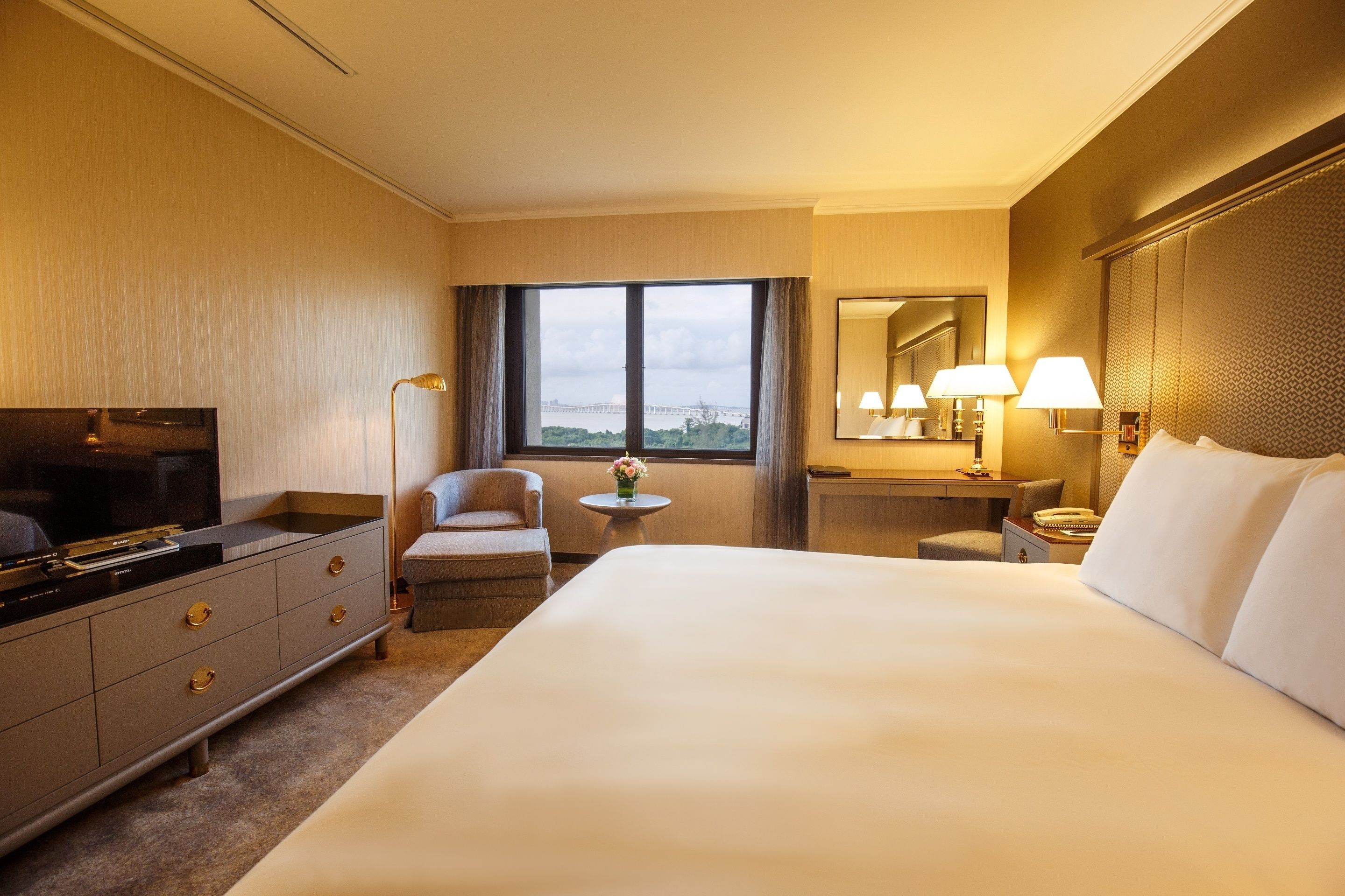A hotel room with a large bed, a desk with a lamp, a chair, a TV, and a view of a bridge.