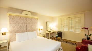 Hotelkamer met een groot bed, een bureau, een stoel, een klein tafeltje met een lamp en een raam met shutters.