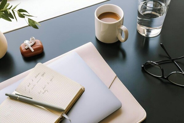 Un carnet rempli de notes manuscrites, un stylo, une paire de lunettes, un ordinateur portable, une tasse de café, un verre d’eau et une plante, le tout posé sur une table.