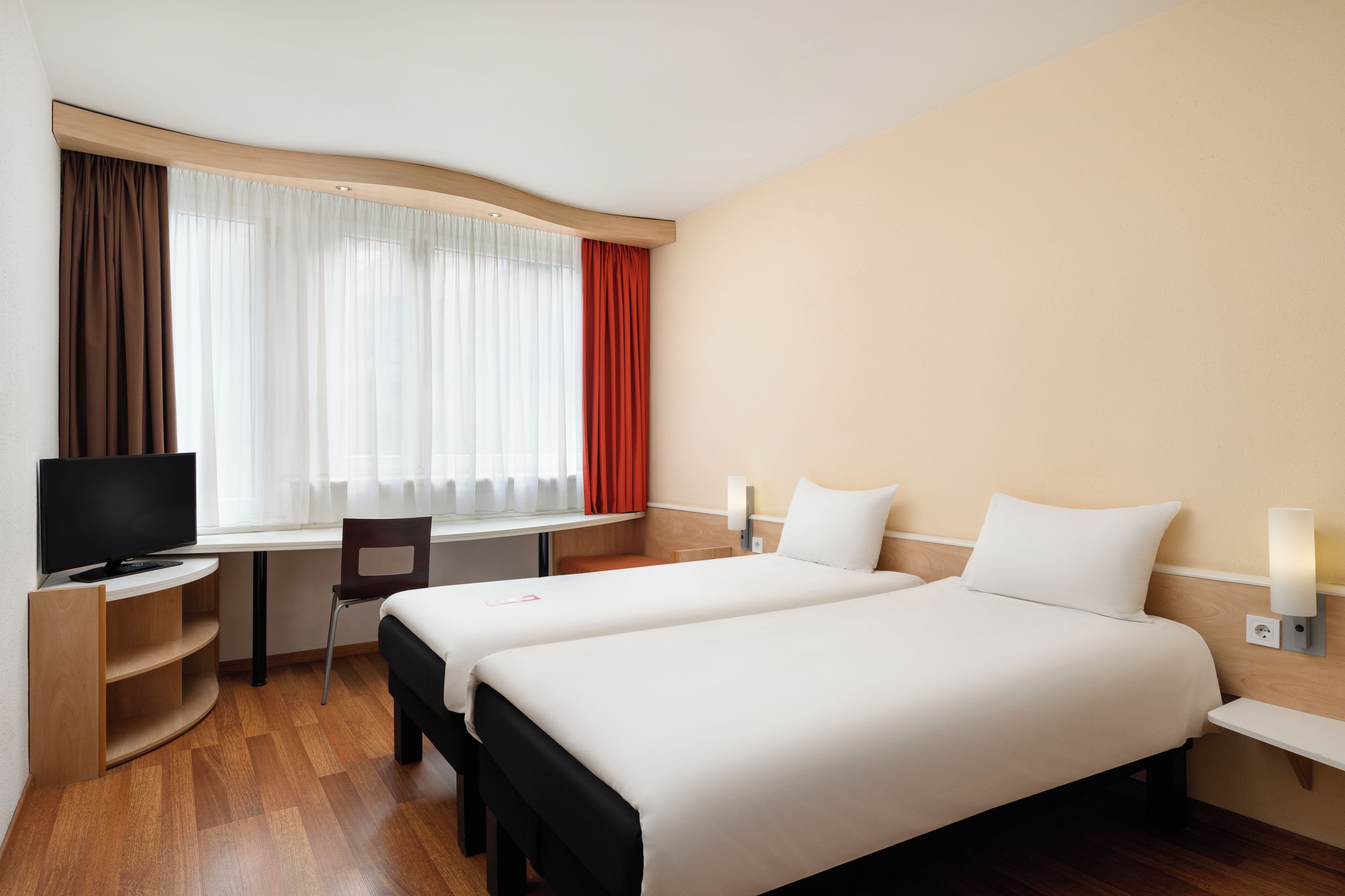 Een hotelkamer met een bed, bureau, stoel, televisie en een raam met gordijnen.