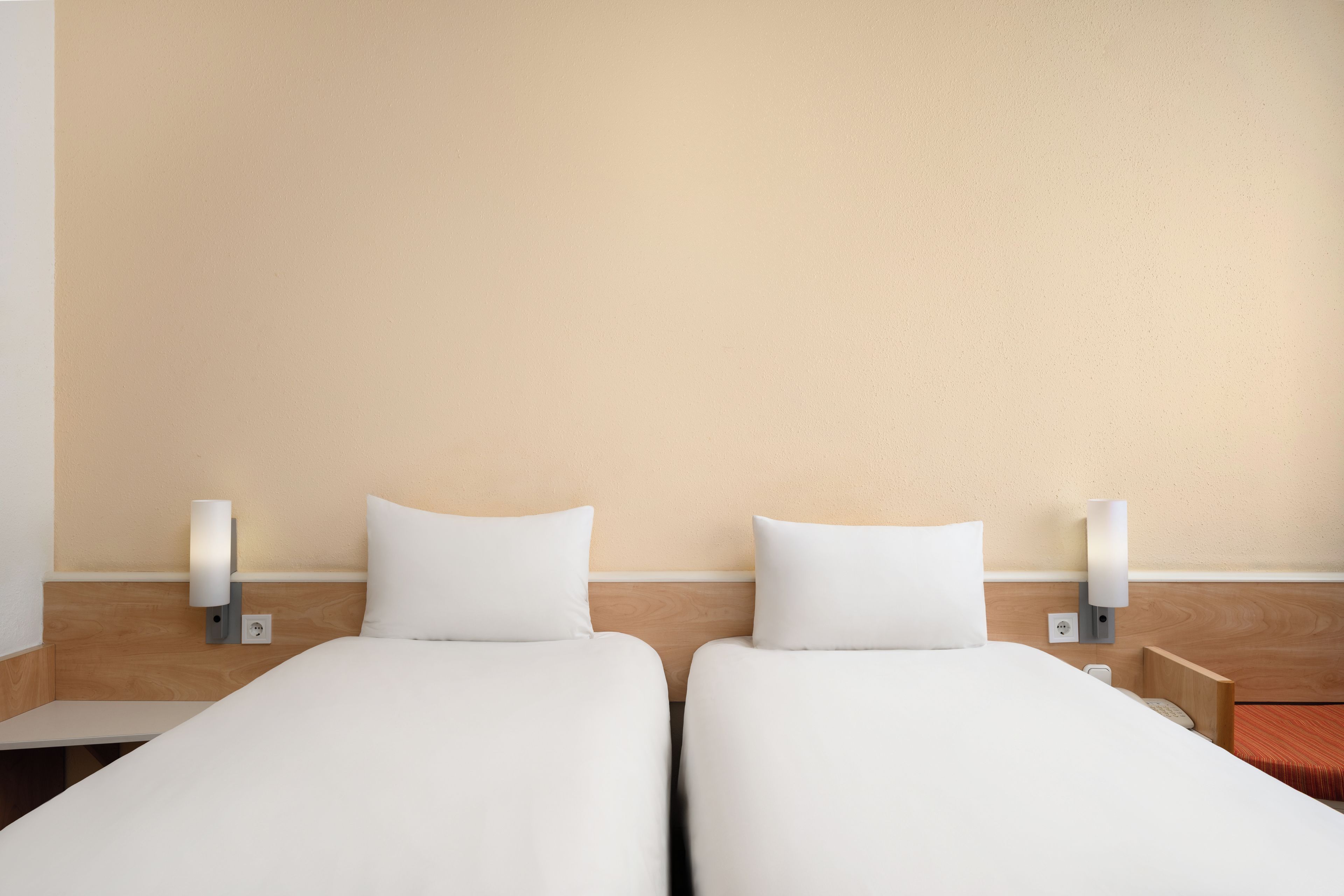 A hotel room with two single beds, each with a white mattress and pillow, flanked by nightstands with wall-mounted lamps.