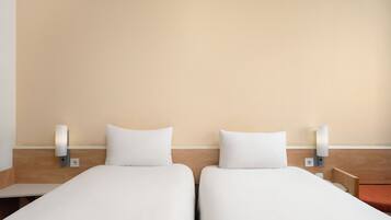 A hotel room with two single beds, each with a white mattress and pillow, flanked by nightstands with wall-mounted lamps.