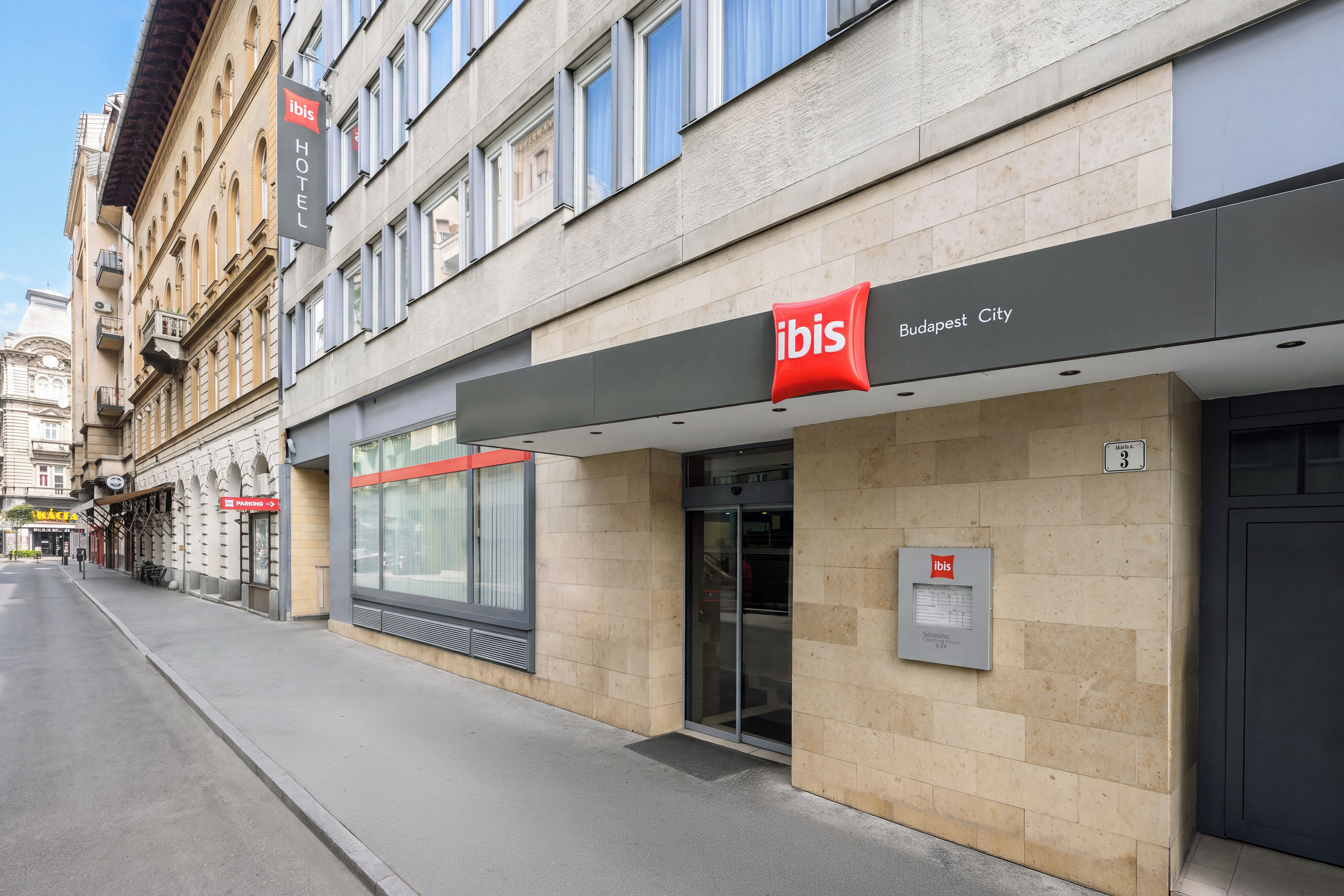 Entrada de un hotel con fachada moderna, un cartel claro que indica 'ibis Budapest City' y un letrero de estacionamiento visible.