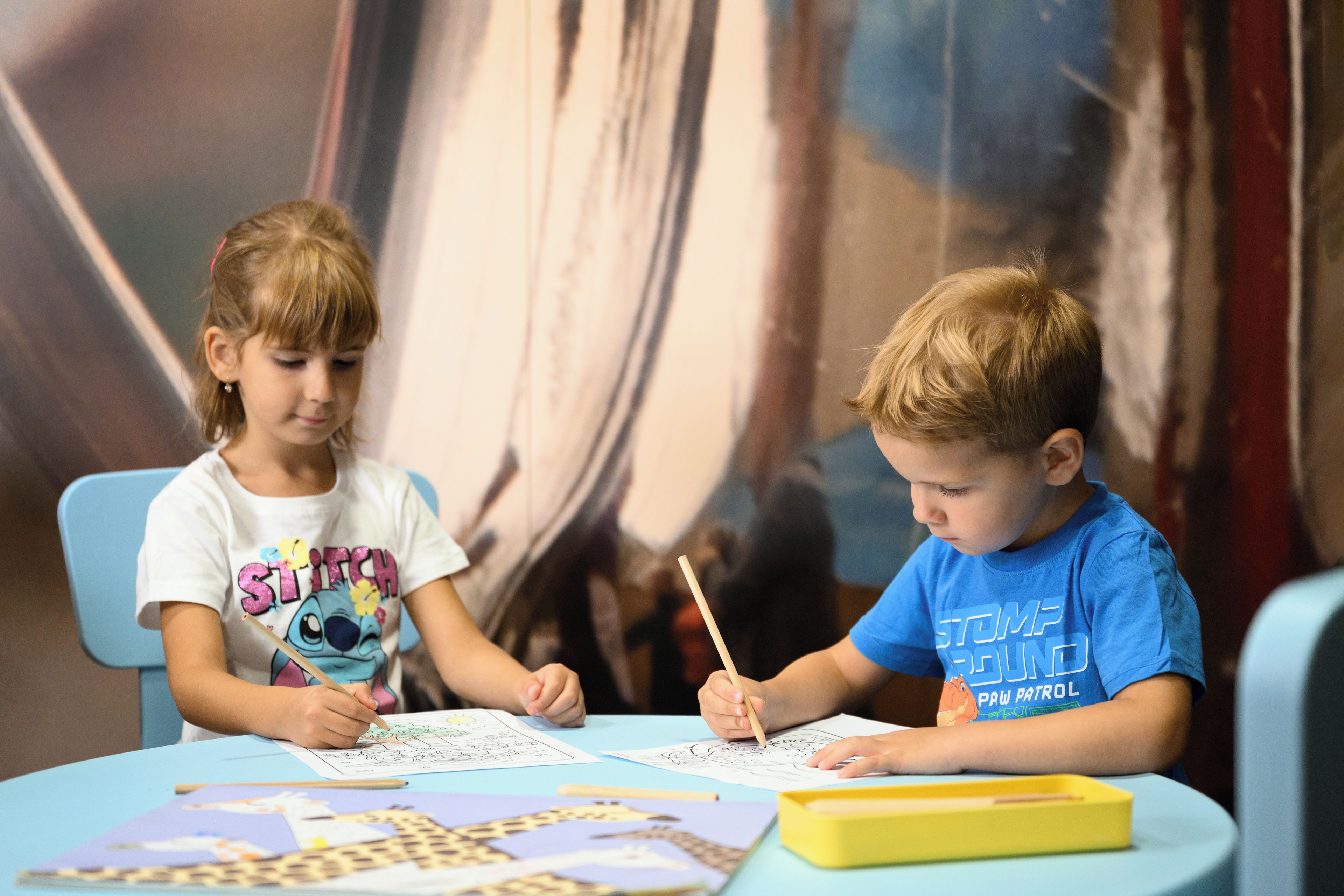 Two children seated at a table, engaged in drawing activities with pencils and paper.