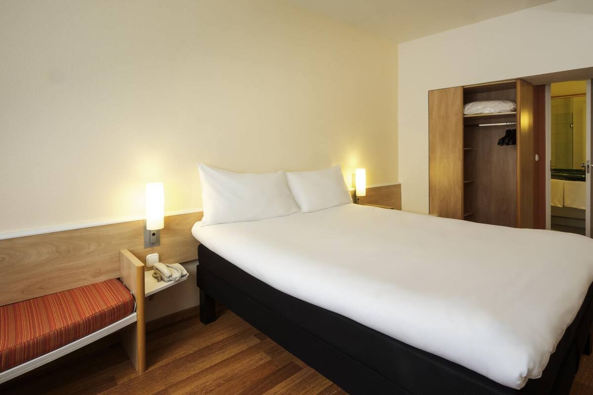 Una habitación de hotel con una cama, mesitas de noche, un armario de madera y un banco.