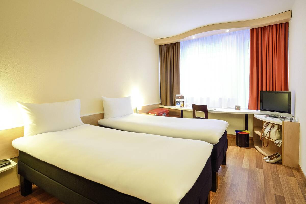 Habitación de hotel con dos camas, un escritorio con computadora, una silla, un televisor y un ventanal con cortinas.