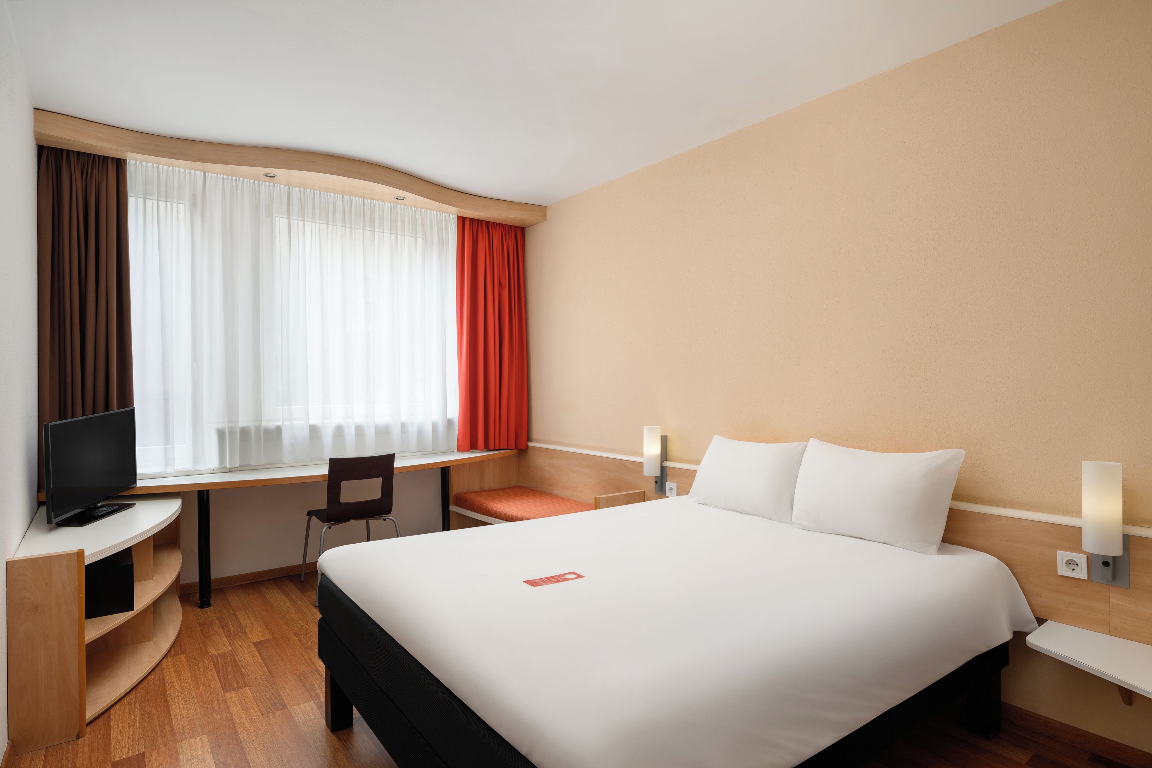 A hotel room with a bed, a desk, a chair, a television, and a window with curtains.