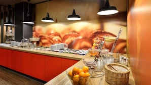 A buffet setup with various food items, a fruit bowl, and a wall with a large image. - ibis Budapest City (Budapest)