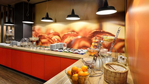 A buffet setup with various food items, a fruit bowl, and a wall with a large image. - ibis Budapest City (Budapest)