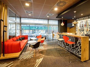A modern interior with a red sofa, multiple chairs, a bar area, and large windows. - ibis Budapest City (Budapest)
