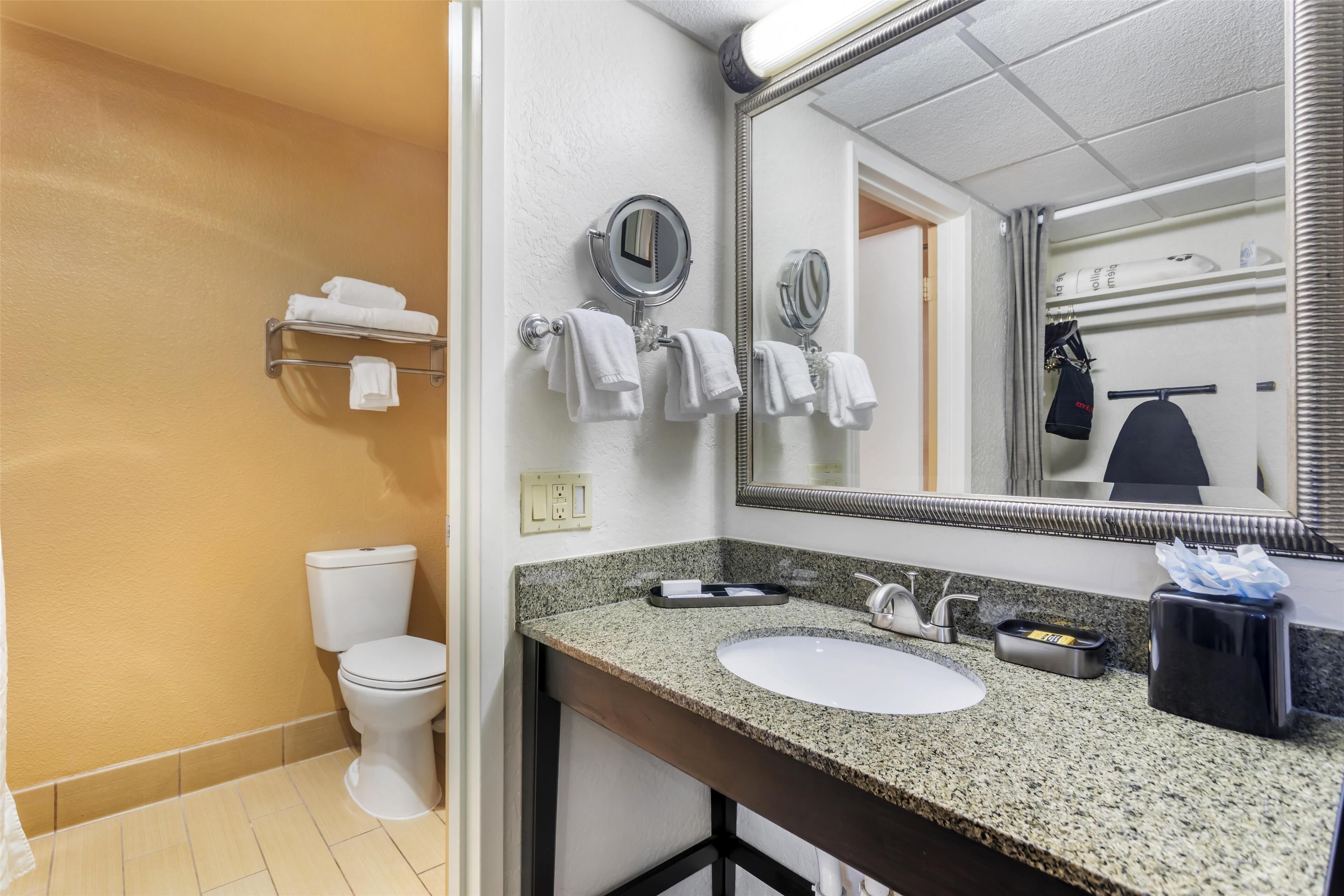 A bathroom with a white toilet, a sink with a granite countertop, a mirror, and a towel rack with white towels.
