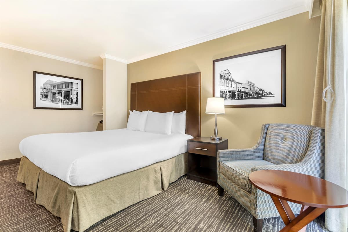 A hotel room with a large bed, a nightstand, a chair, a table, and two framed pictures on the wall.