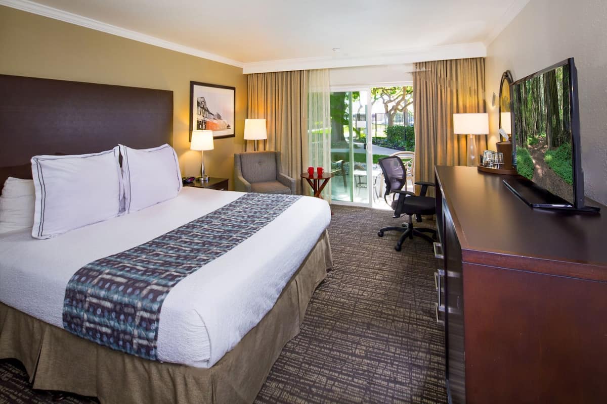 A hotel room with a large bed, two bedside tables with lamps, a desk with a TV, and a view of the outdoors through a sliding glass door.