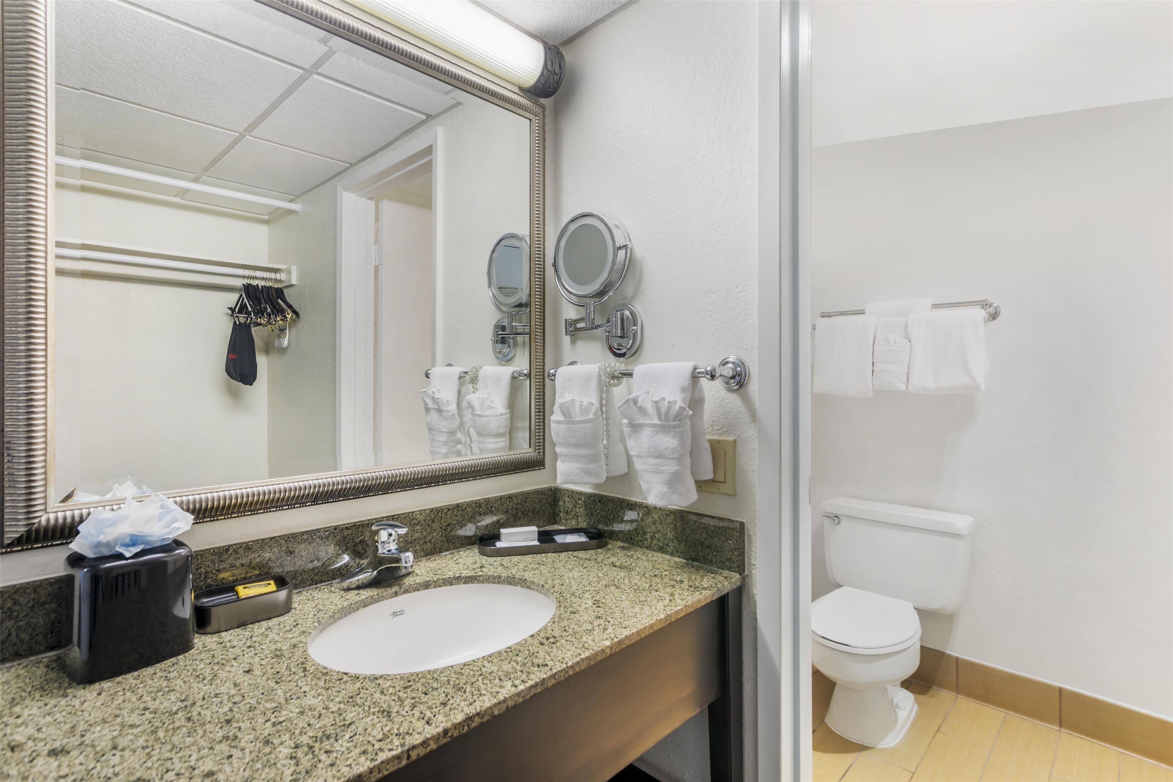 A bathroom with a granite countertop, a white sink, a large mirror, two round mirrors, a towel rack with white towels, a toilet, and a trash bin.