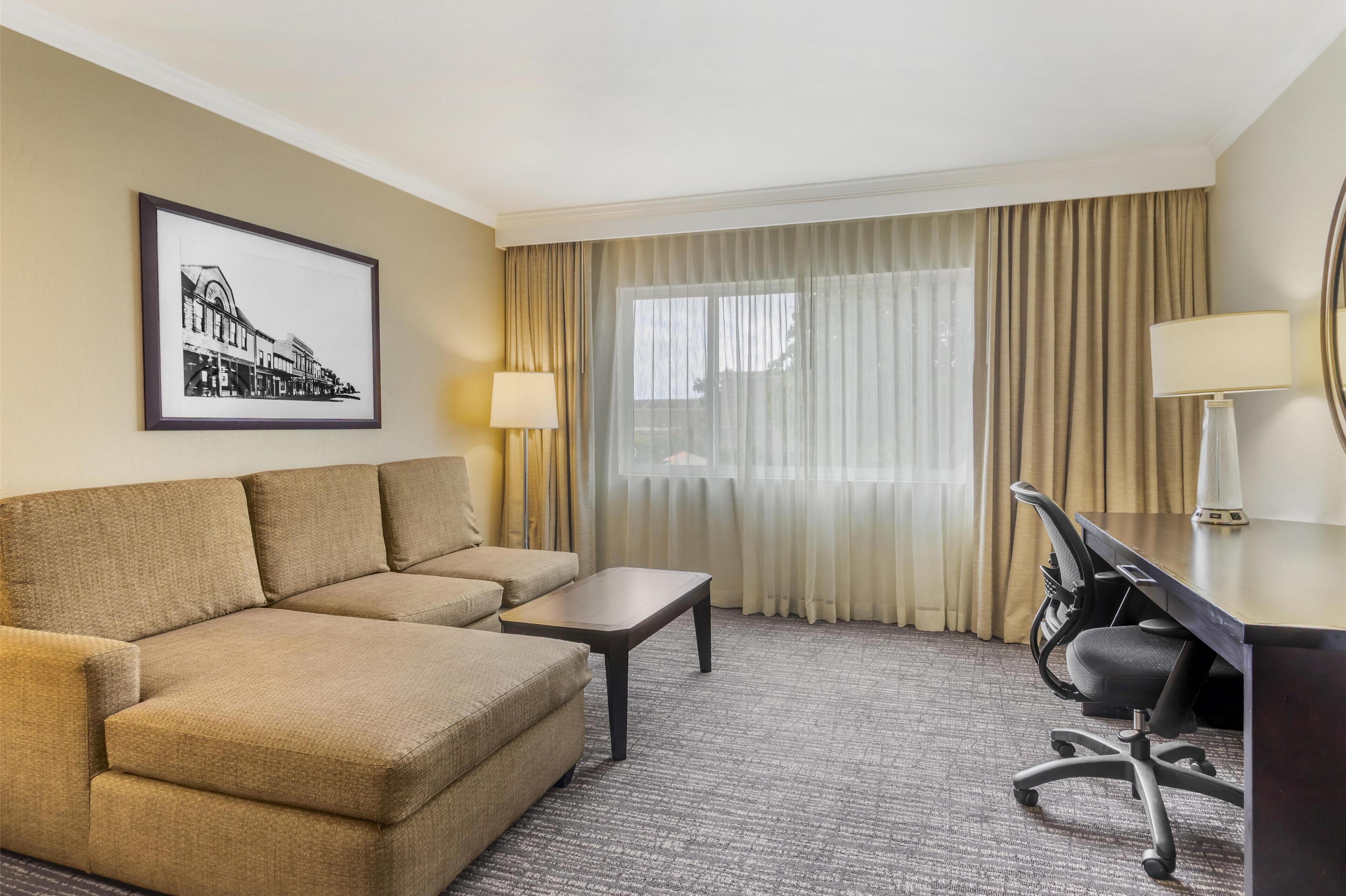 A hotel room with a sofa, a small table, a desk with a chair, a lamp, a large window with curtains, and a framed picture on the wall.
