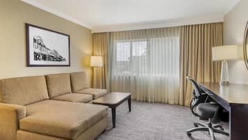 A hotel room with a sofa, a small table, a desk with a chair, a lamp, a large window with curtains, and a framed picture on the wall.