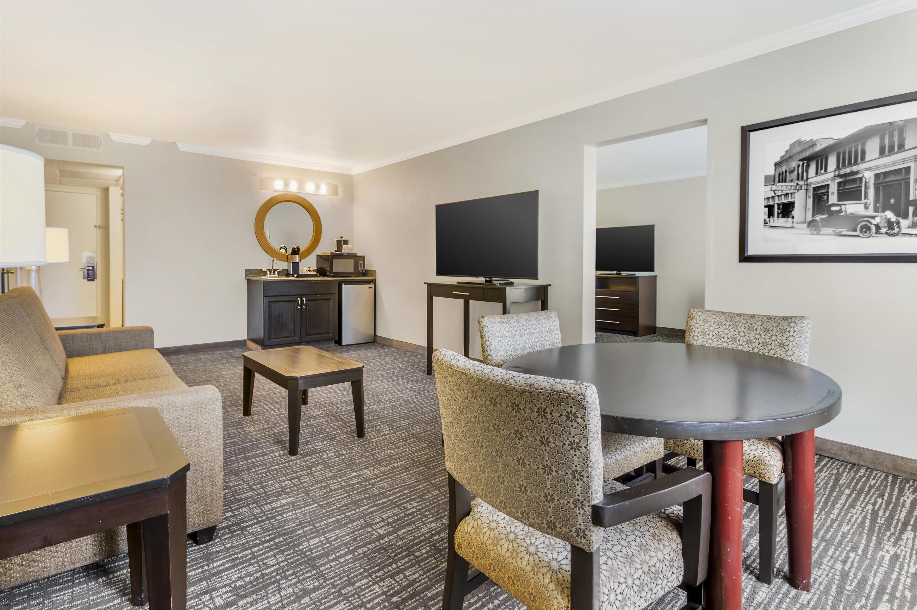 A hotel room with a sofa, a small table, a round dining table with chairs, a TV, a mirror, a framed picture on the wall, and a kitchenette.