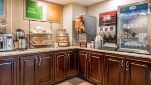 A hotel kitchen with a coffee station, a fruit bowl, and a refrigerator stocked with beverages.
