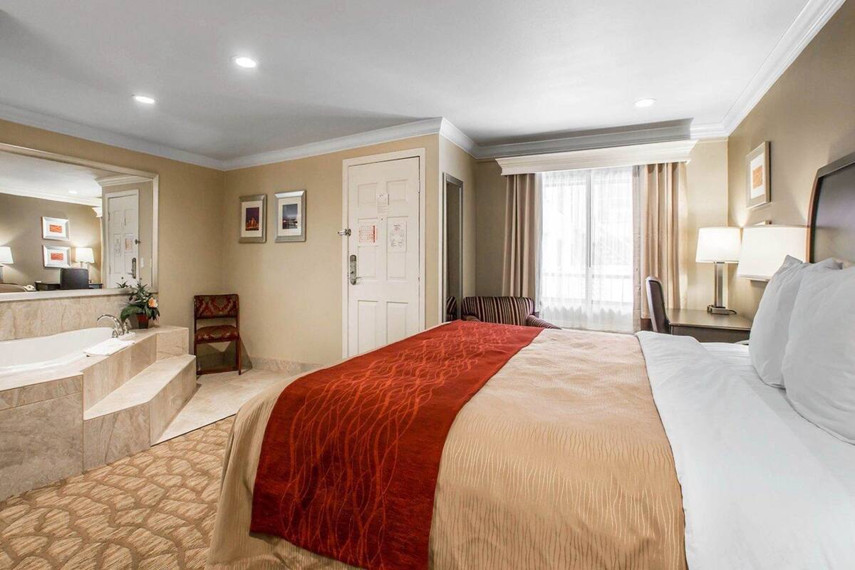 A hotel room with a bed, a desk, a chair, a bathroom with a sink, and a mirror.