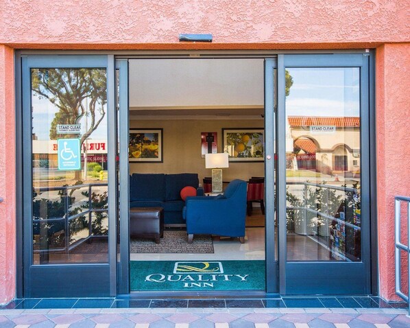 Entrada de hotel con puerta corredera, felpudo verde con el nombre del hotel, sofá azul en el interior y señal de acceso para sillas de ruedas en el exterior.
