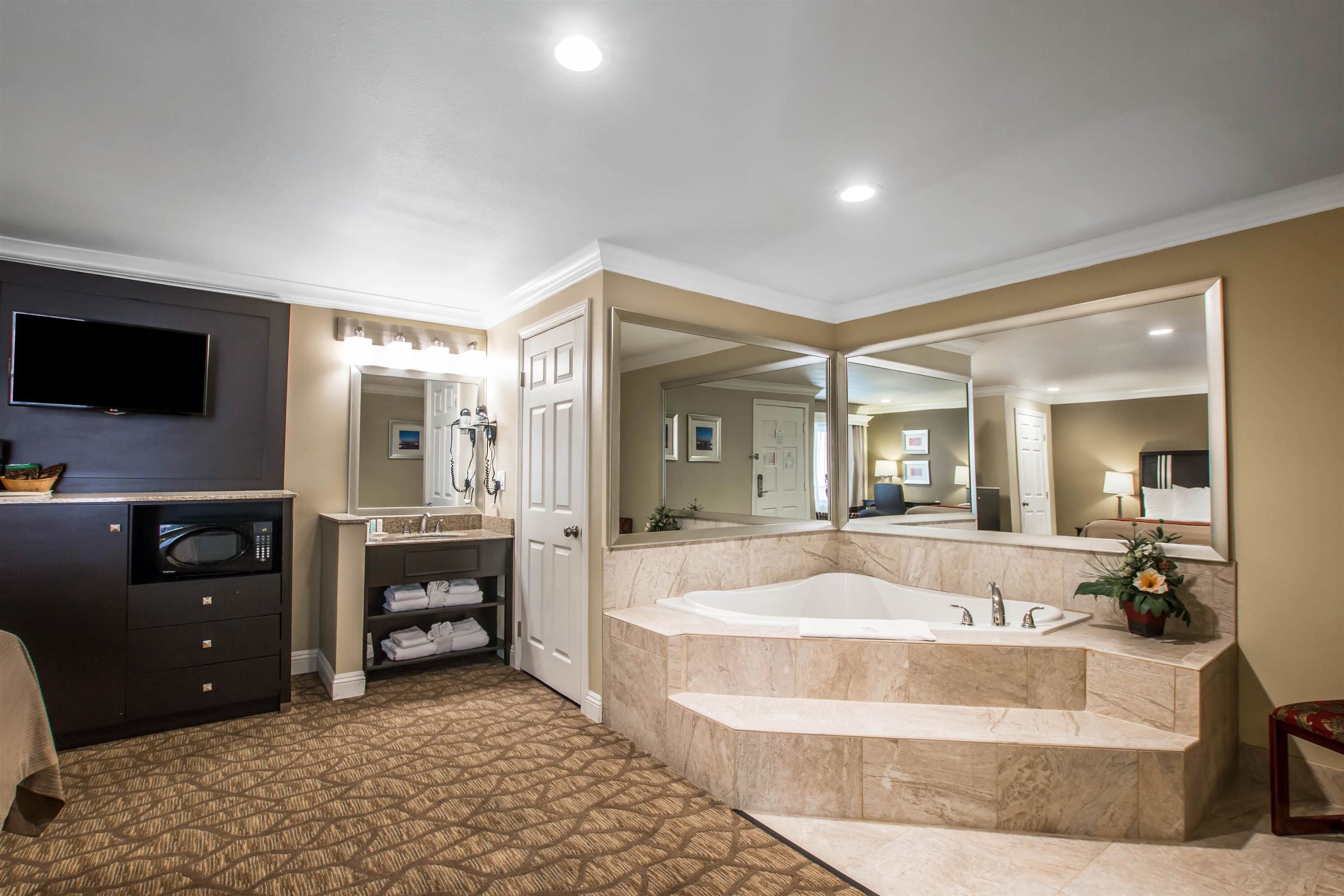 A modern bathroom with a large bathtub, a vanity with a mirror, a microwave, and a television.