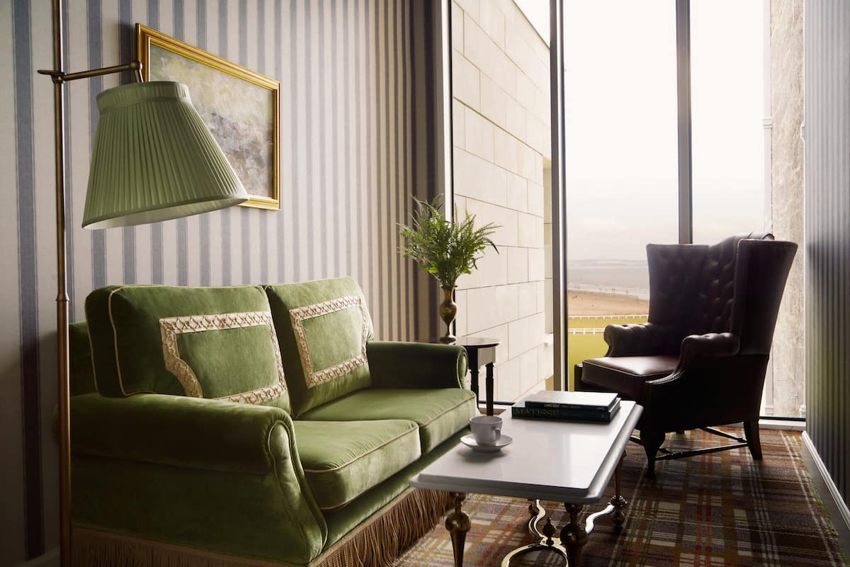 A green sofa with patterned pillows, a white table, a dark armchair, a floor lamp, and a framed artwork on the wall.