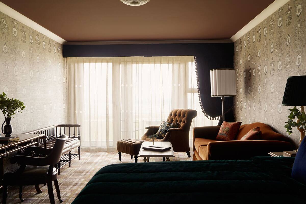 A hotel room with a large bed, a sitting area with a sofa and armchair, a desk with a chair, and a large window with sheer curtains.