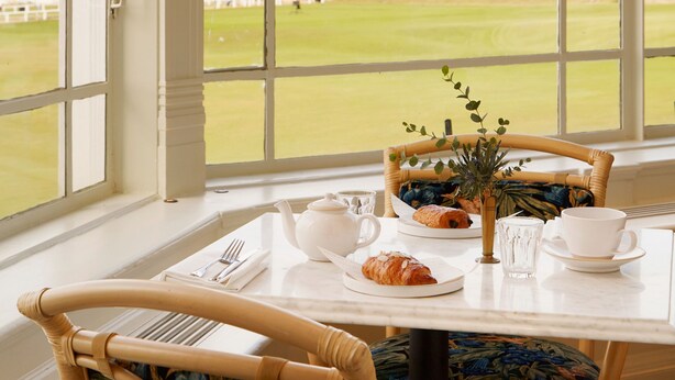 A dining area with a table set for breakfast, a wicker chair, and a view of a large building through large windows.