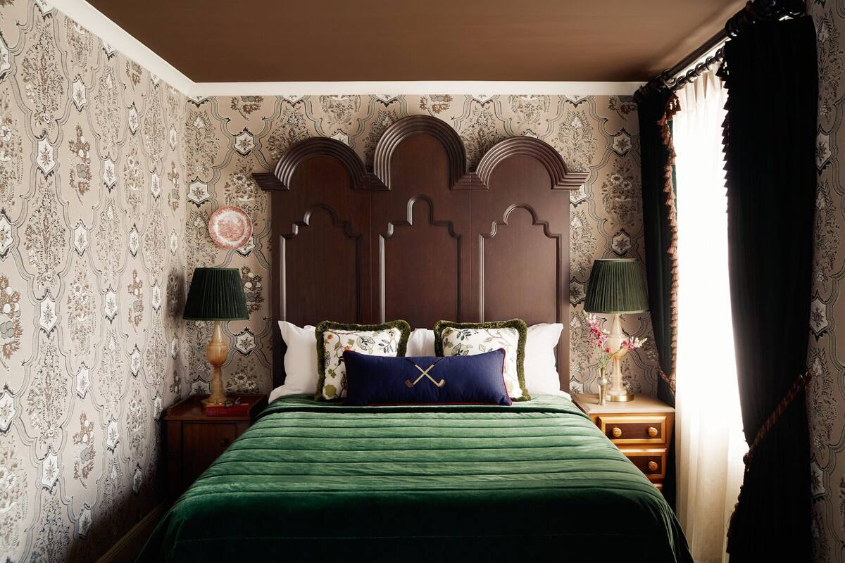 A bedroom with a green bedspread, a decorative headboard, patterned wallpaper, and two bedside tables with lamps.