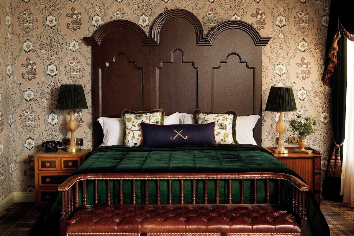 A traditional bedroom with a large wooden headboard, a green bedspread, and matching green curtains. There are two bedside tables with lamps and a small bedside table with a telephone.