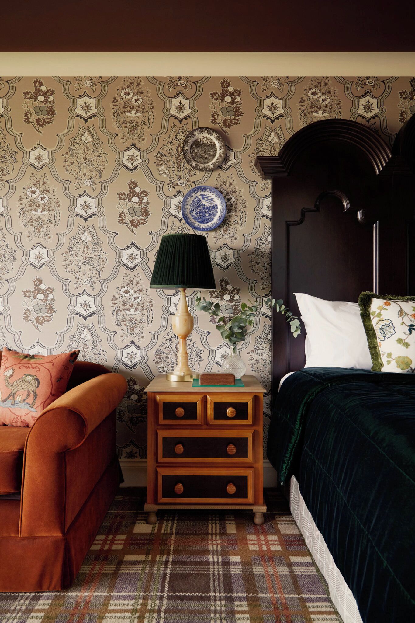 A bedroom with a patterned wallpaper, a wooden nightstand with drawers, a green lamp, a decorative plate, a dark headboard, and a plaid rug.